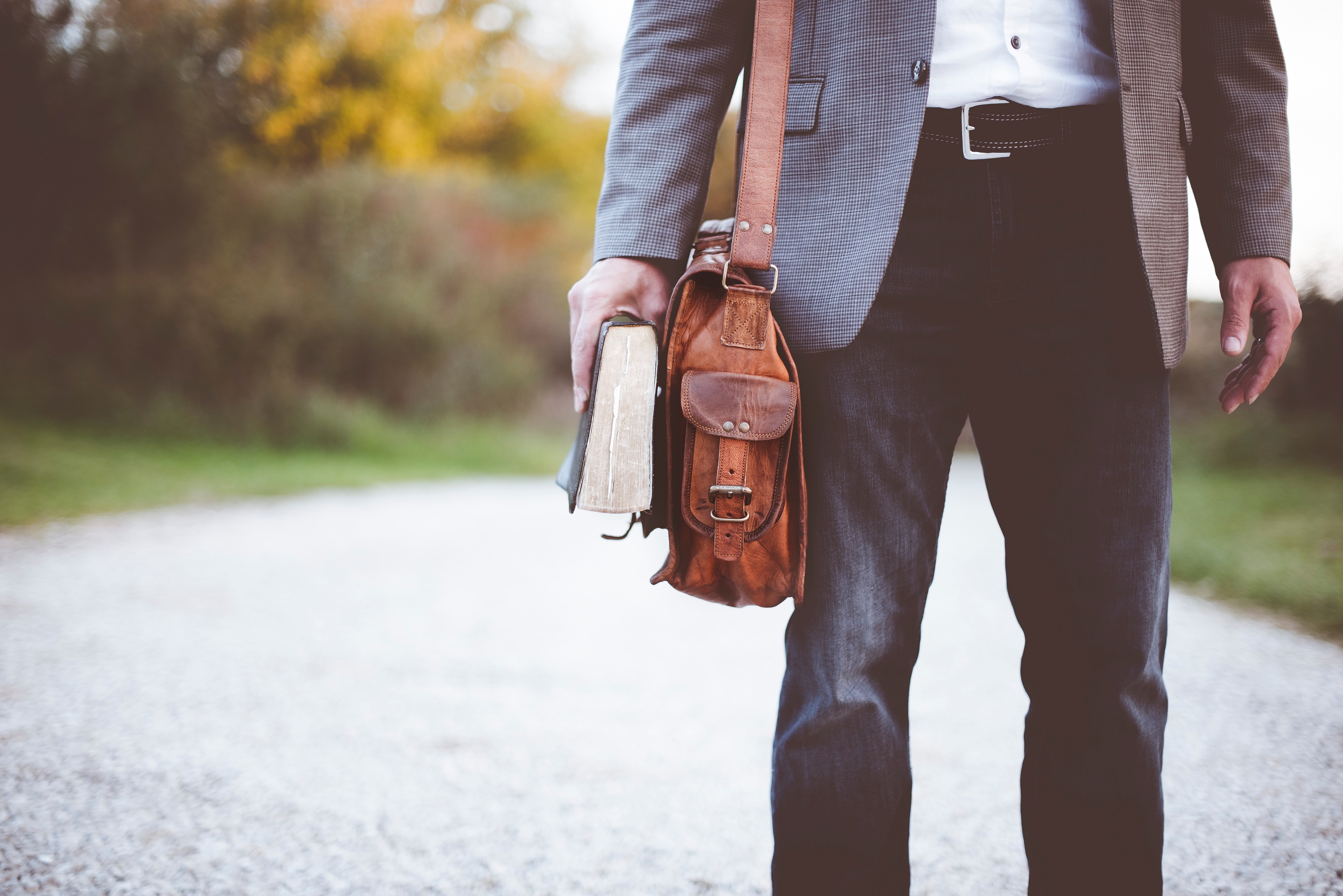 A man can be partially seen carrying a brown leather shoulder bag and a thick book.